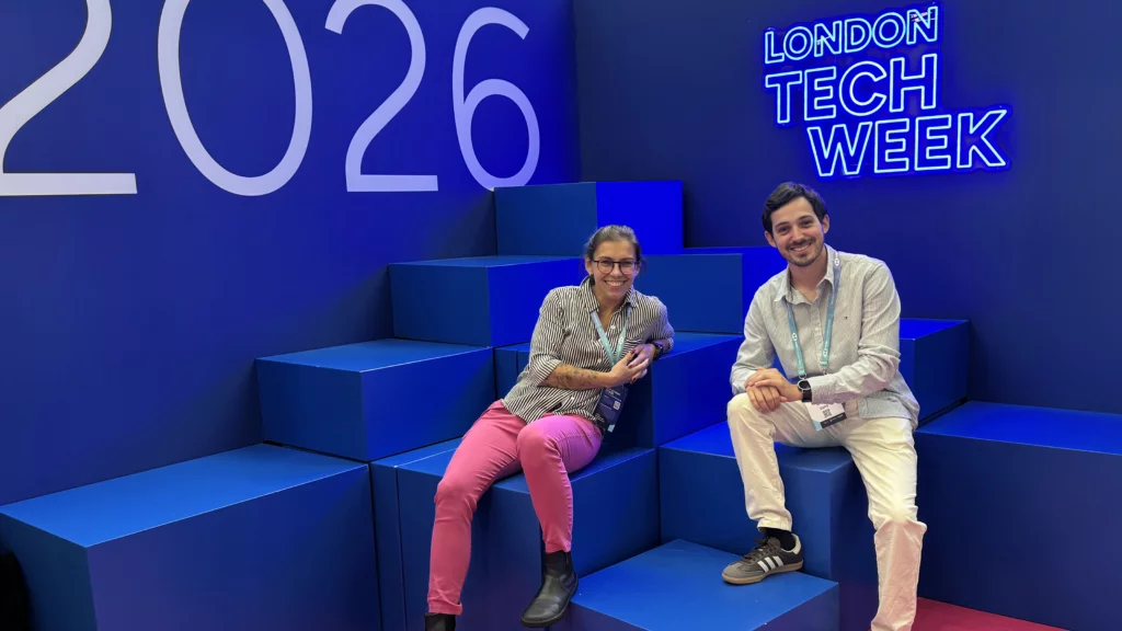 Two attendees smiling while sitting on blue steps at London Tech Week 2026, with a neon sign in the background.