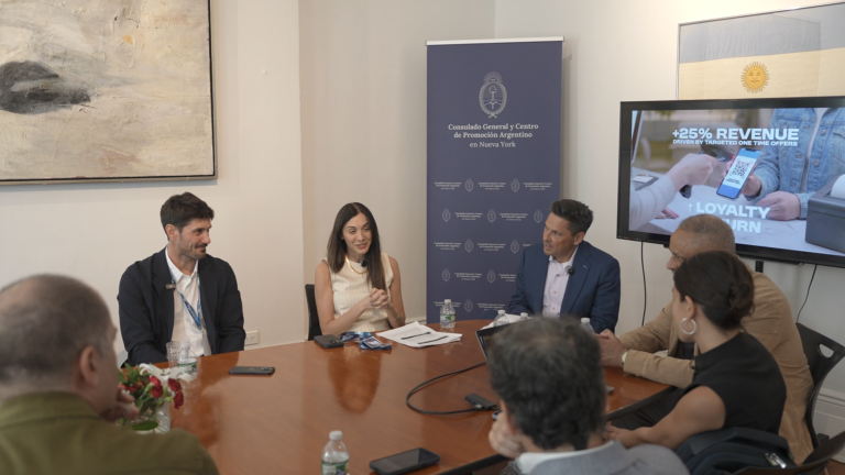 Group discussion at a conference table featuring several participants, with a presentation about business strategies in the background.